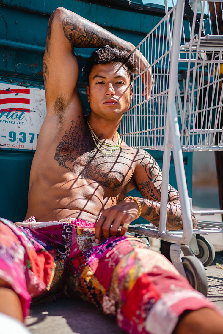 Topless, muscular man with tattoos sits against a blue dumpster and shopping cart.