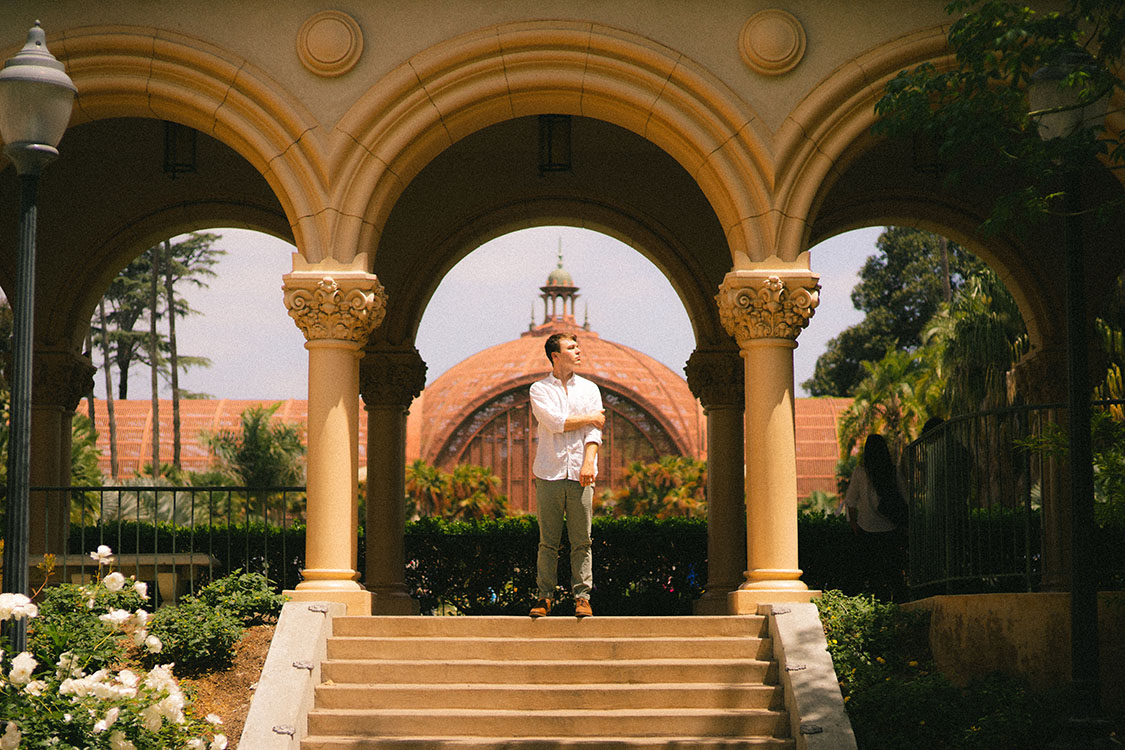 Man in white button-up shirt stands under Spanish archway at top of stairs.