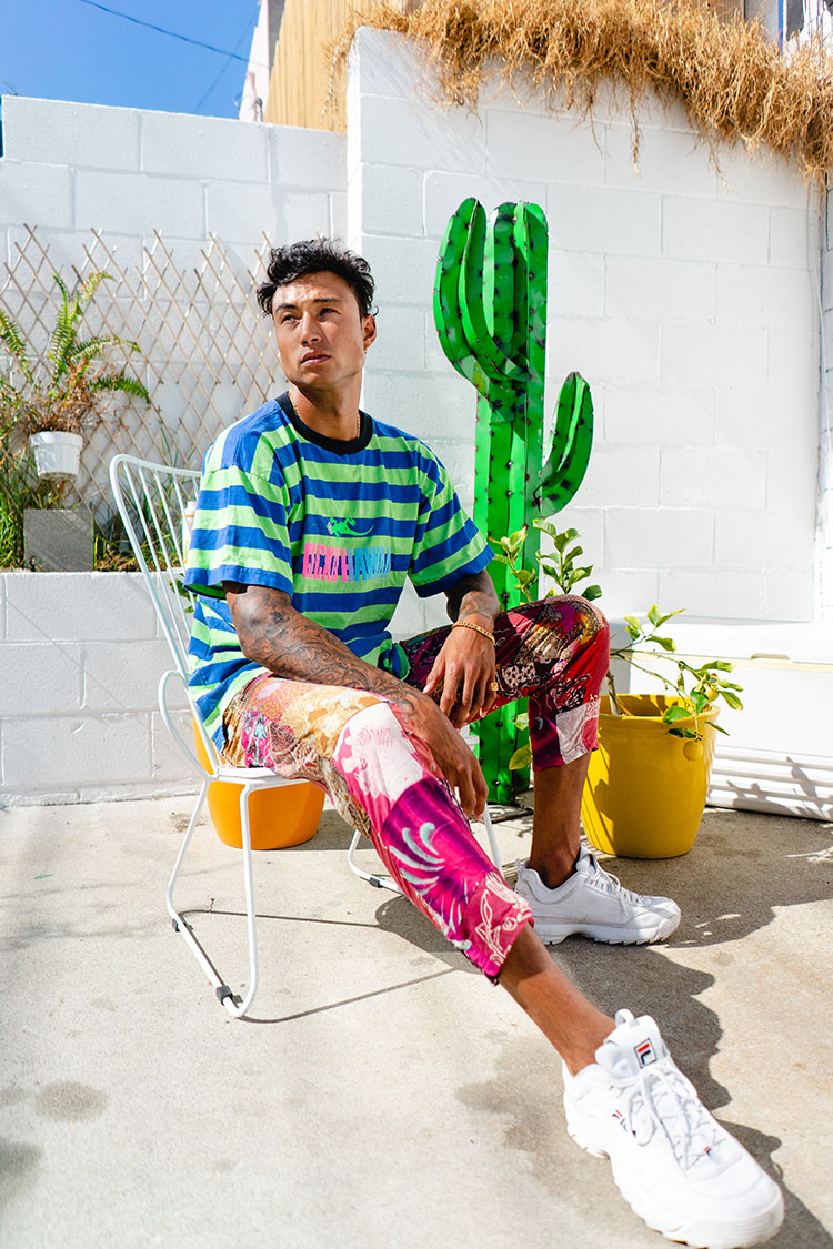 Man in colorful clothes sits on a white outside with a green cactus sculpture behind him.
