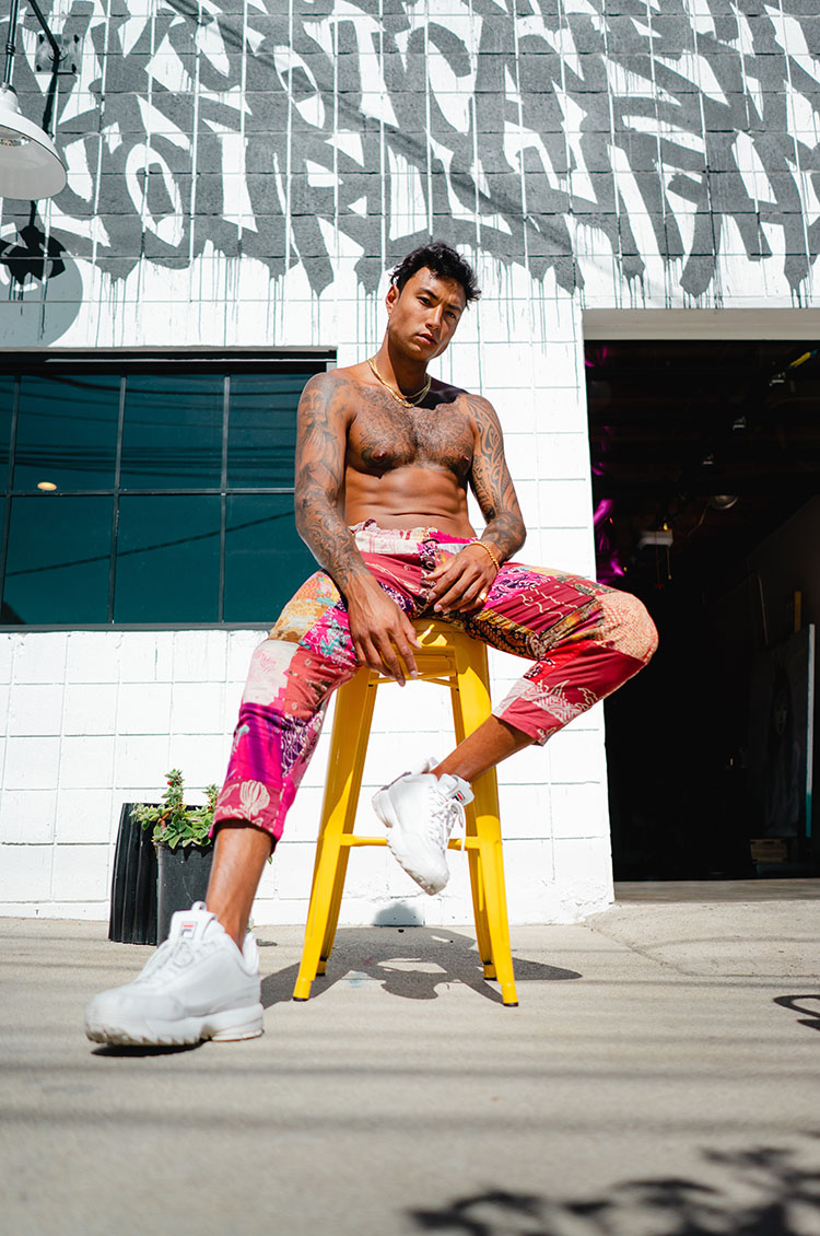 Topless, muscular man with tattoos sits outside on yellow stool in front of a geometric-painted black-and-white brick wall.