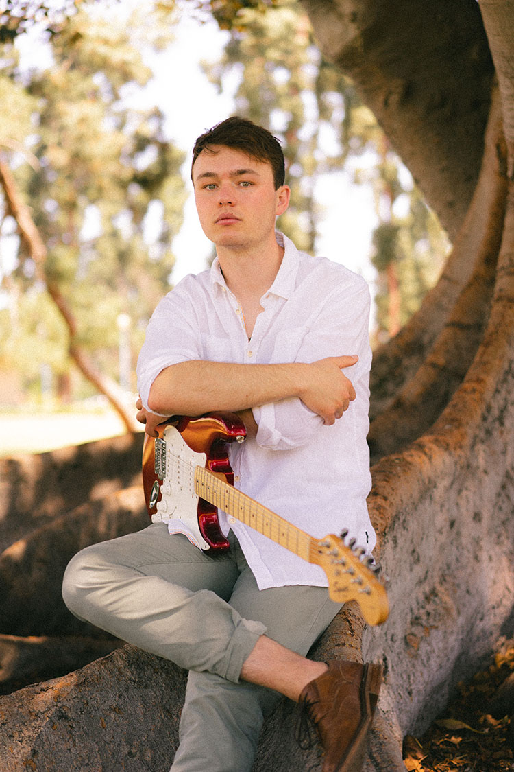 Man in white button-up shirt sits on a Moreton Bay fig tree with his electric guitar.