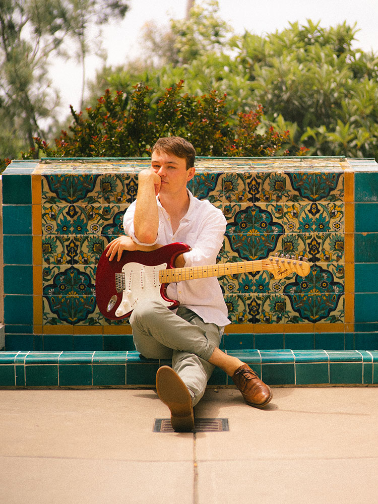 Man in white button-up shirt sits with electric guitar in front of a Spanish tile fountain.