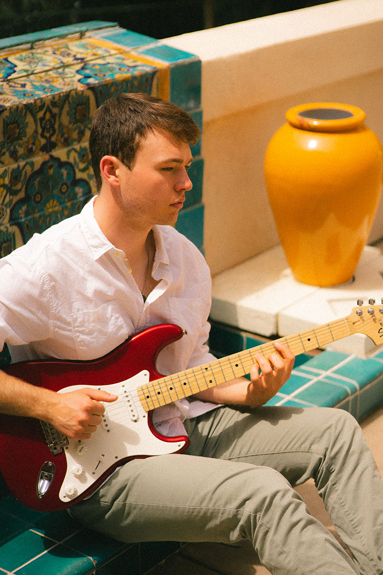 Man in white button-up shirt plays electric guitar in front of a Spanish tile fountain.