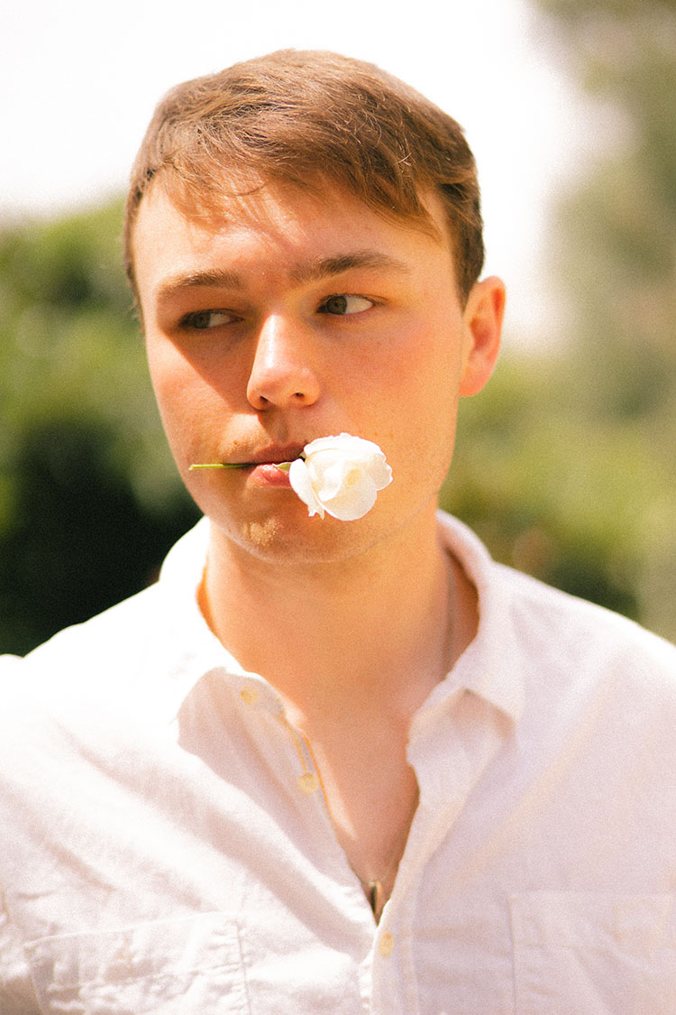Man in white button-up shirt holds a white flower between his lips.