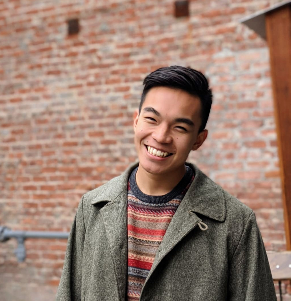 Barrett Lo wearing a red sweater and green coat in front of a red brick wall.
